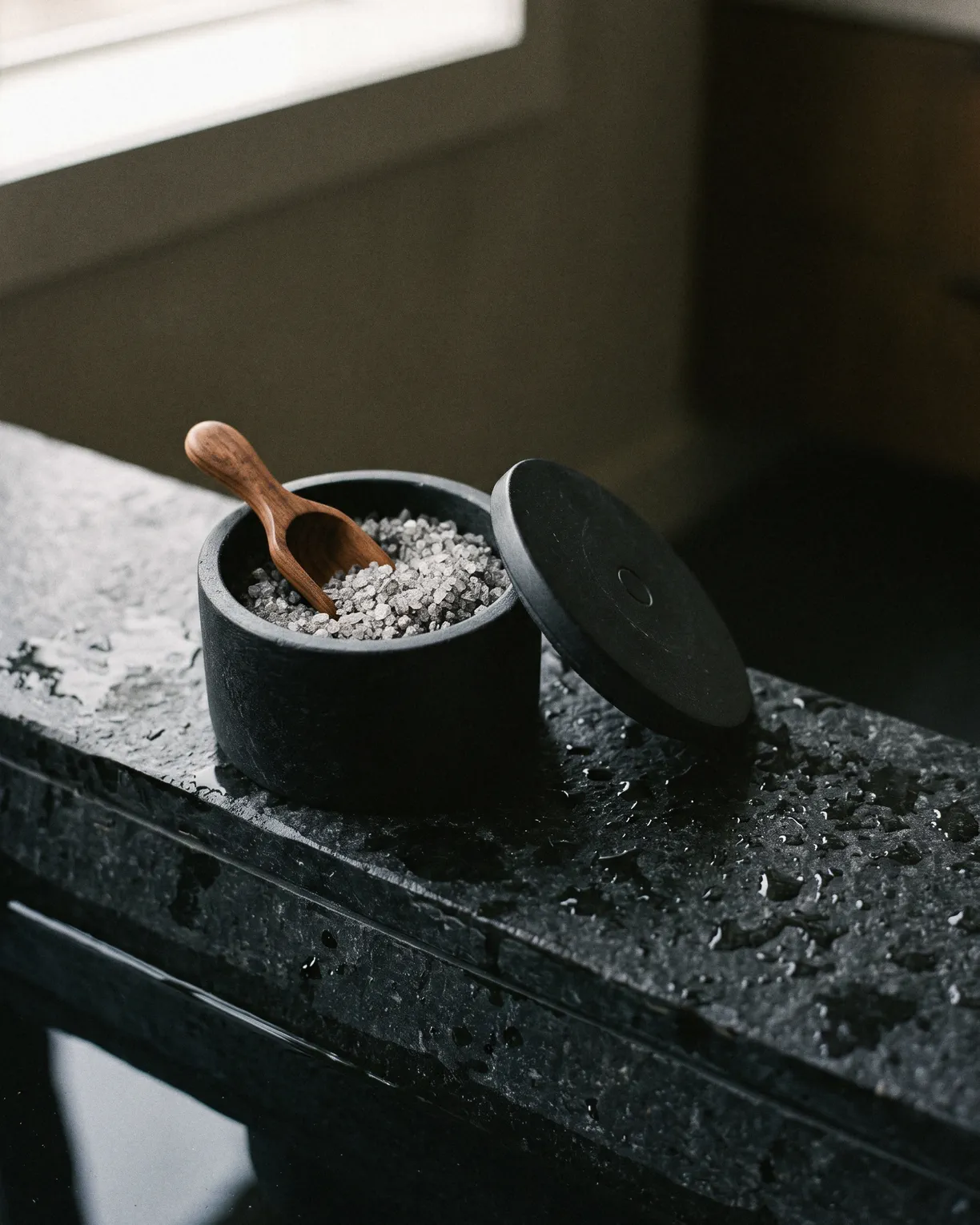 the bath soak, grey mineral salts in a black stone jar with a walnut scoop, on wet black basalt
