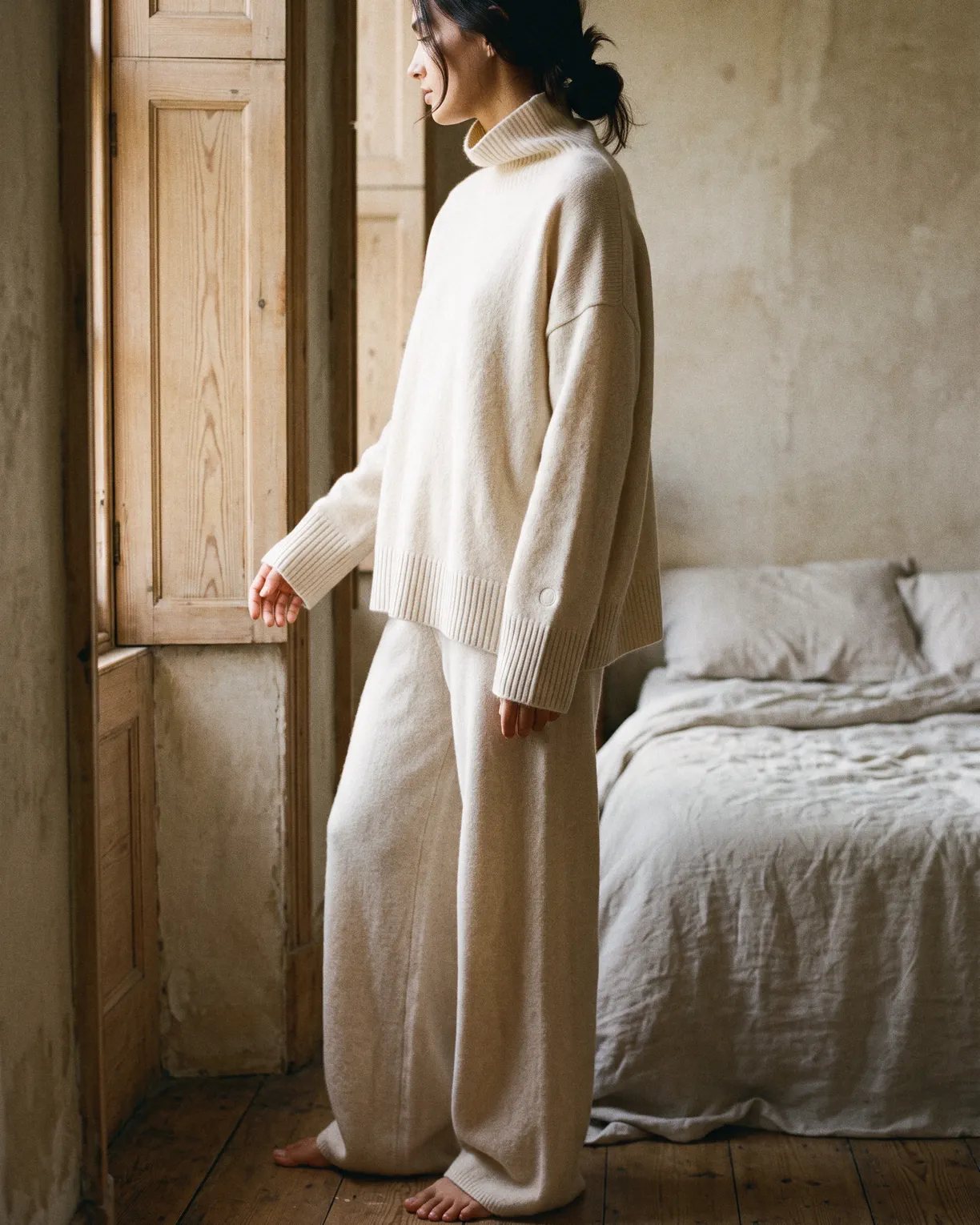 the cashmere set in coconut cream, a loose funnel-neck sweater and wide trousers, worn barefoot by a bedroom window