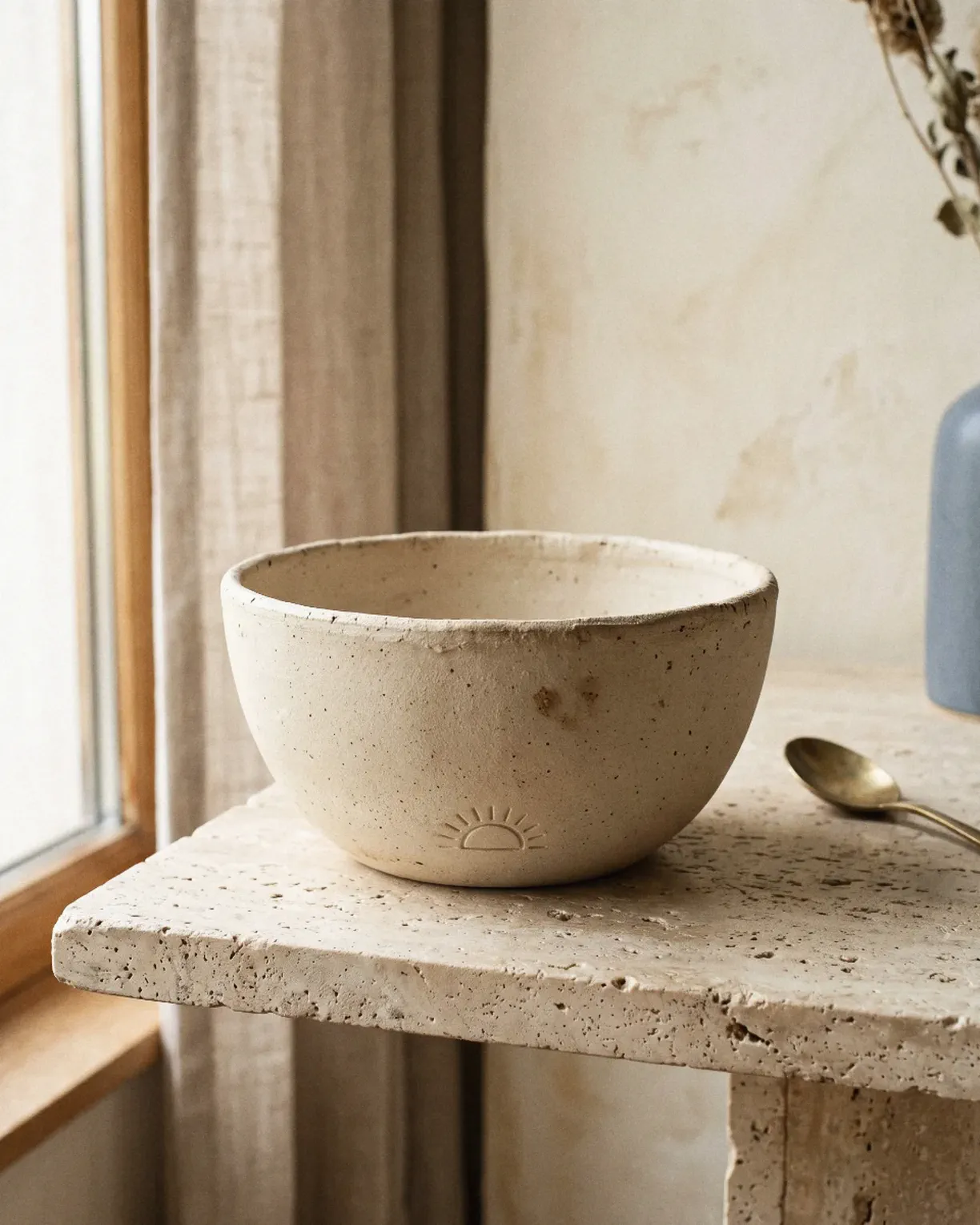the dawn bowl — cream stoneware bowl with a small sun pressed near its base, on a travertine table
