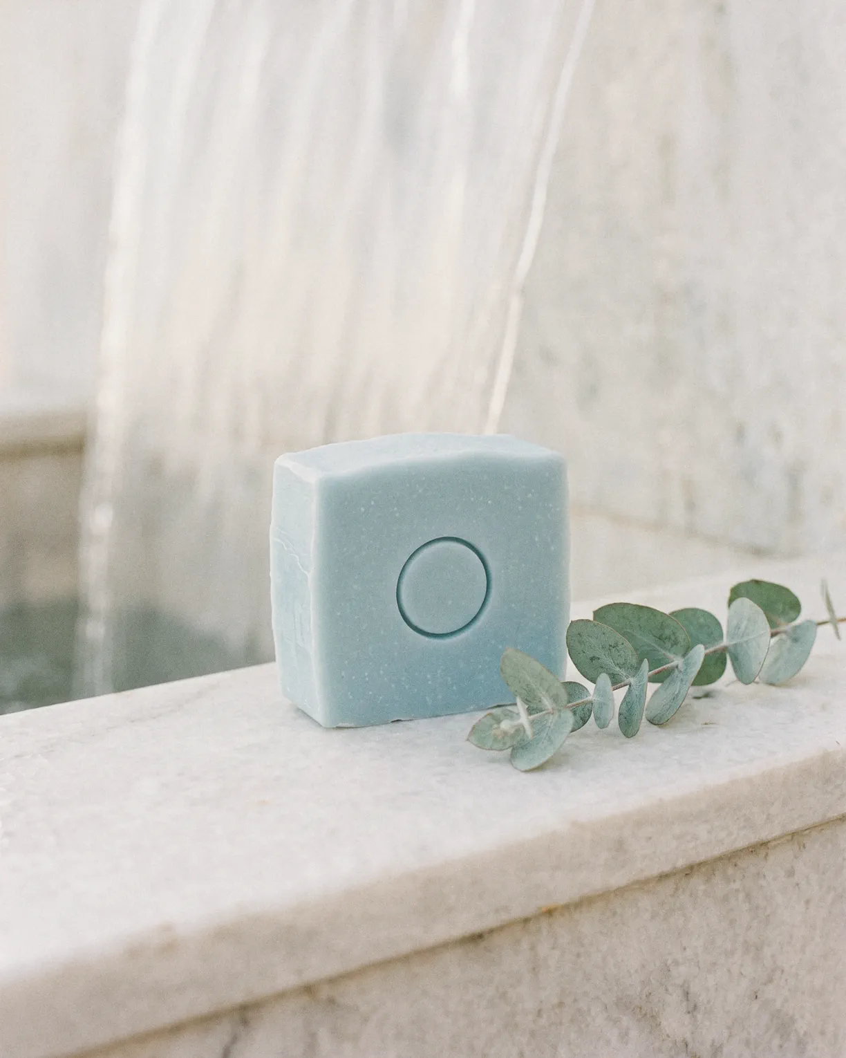 the glacier bar, ice blue soap pressed with a circle, beside a sprig of eucalyptus on white marble under falling water