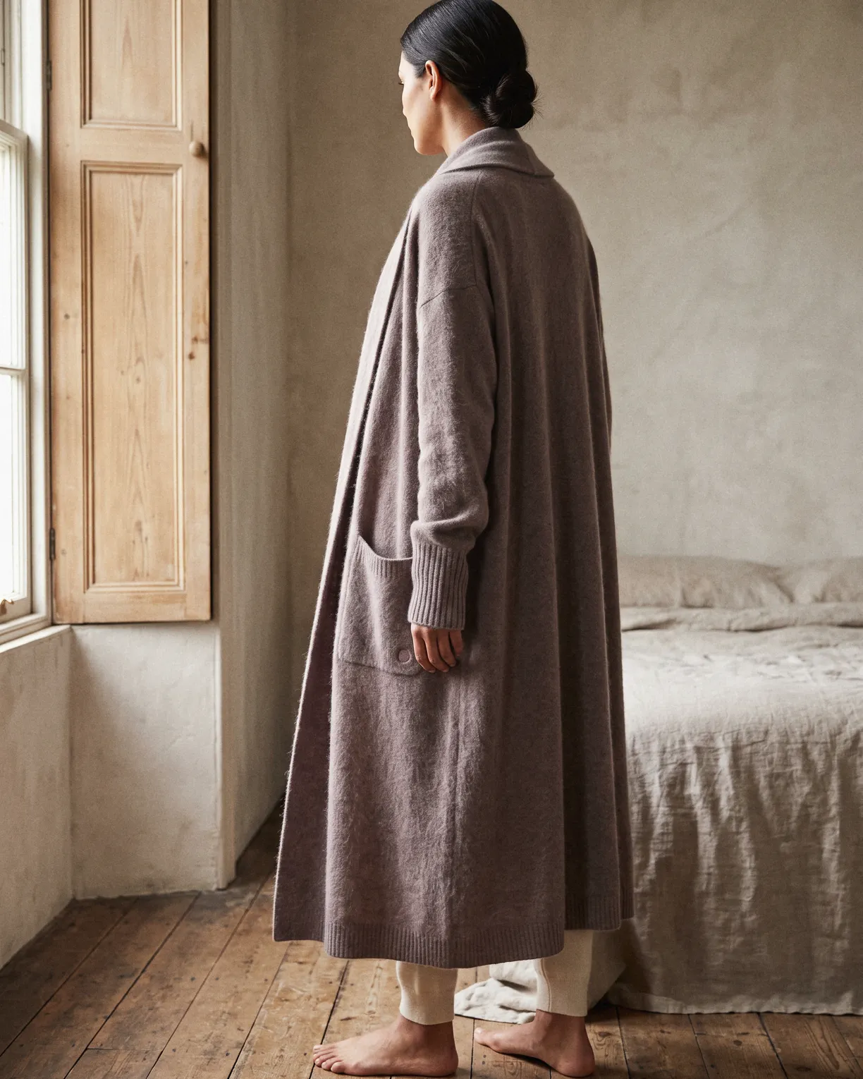 the long cardigan, ankle-length brushed cashmere in a soft taupe, worn open over cream by a bedroom window
