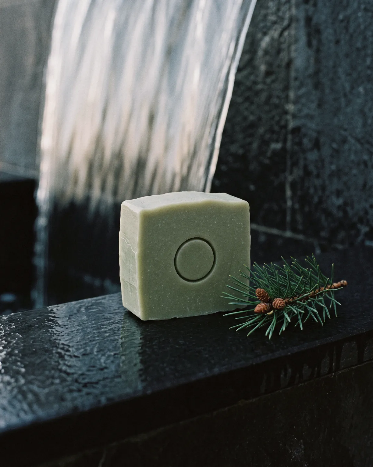 the moss bar, sage green soap pressed with a circle, beside a sprig of pine on wet black basalt under falling water