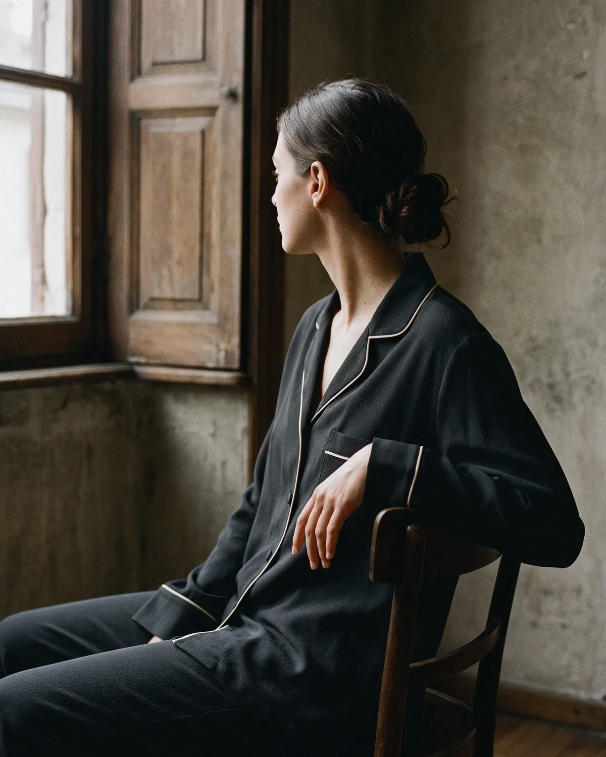 the silk pajamas in black silk crepe piped in cream, worn seated on a wooden chair by a window