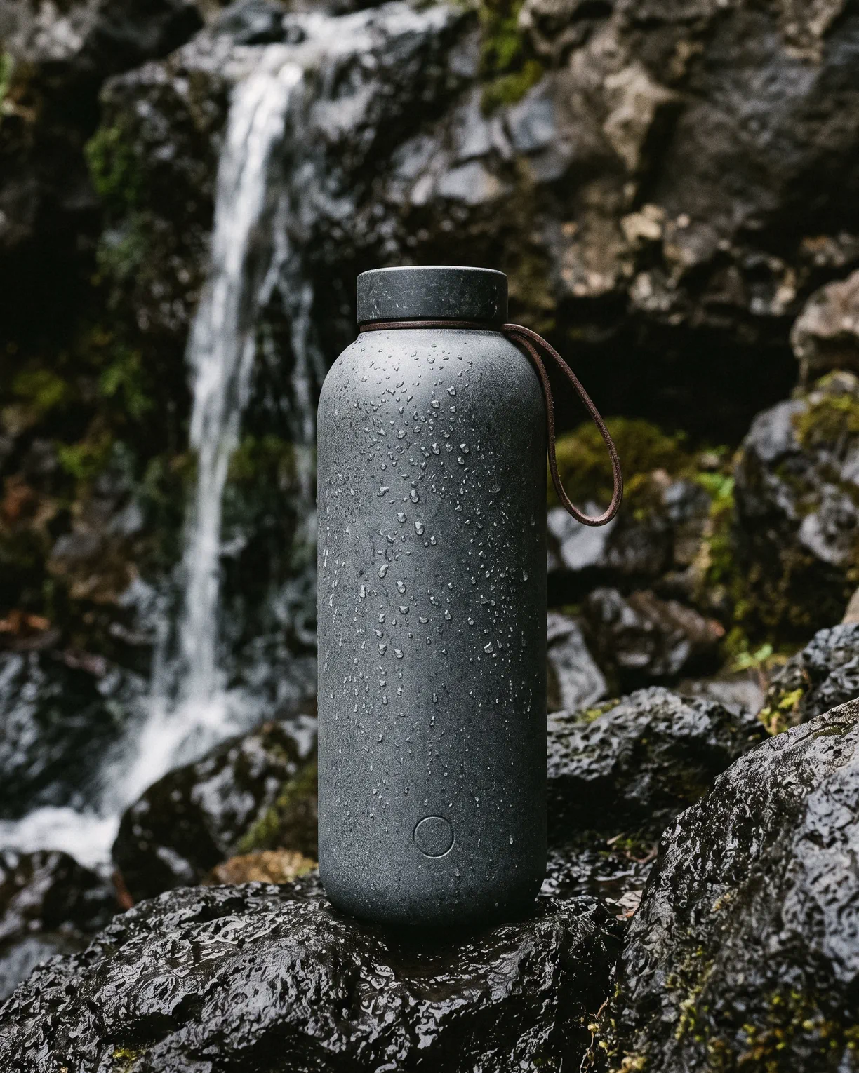 the stone flask, a basalt grey insulated flask with a stone cap and a leather loop, on wet rock by a waterfall