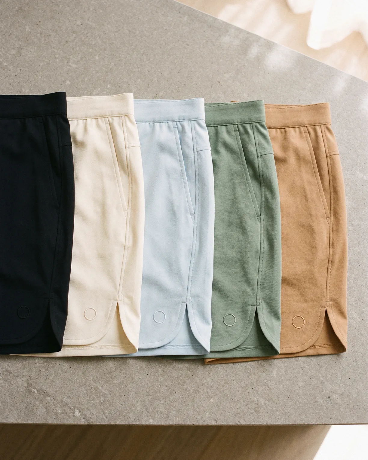 the water shorts in all five colors, black, coconut cream, glacier, moss and dusk, laid side by side on stone